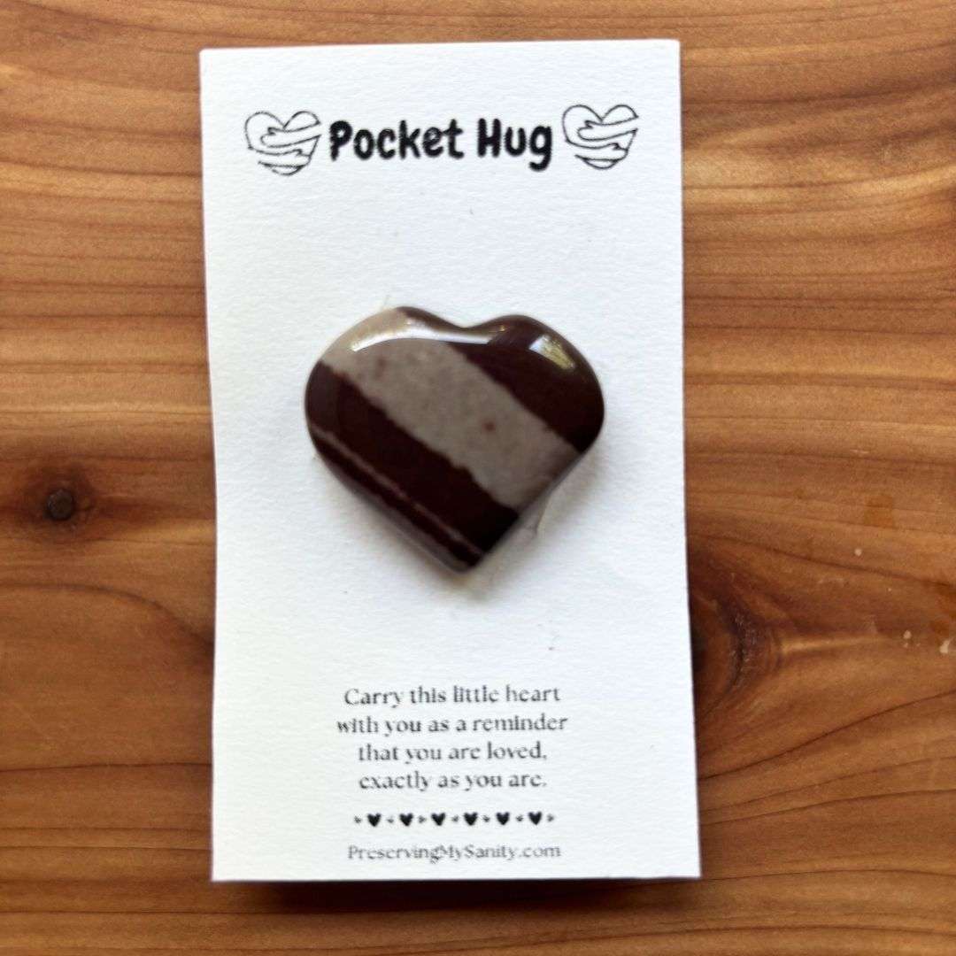 A polished heart-shaped narmada is mounted on a white "Pocket Hug" card and displayed on a warm cedar wood background. The card features the words "Pocket Hug" at the top with heart illustrations and a heartfelt message below: "Carry this little heart with you as a reminder that you are loved, exactly as you are." Small black hearts decorate the bottom above the Preserving My Sanity website, making this a thoughtful keepsake for encouragement, comfort, or gifting.