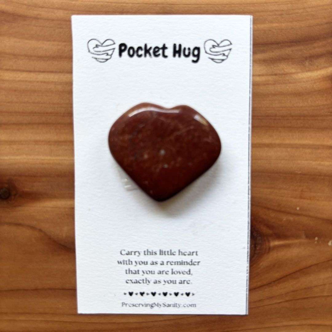 A polished heart-shaped red jasper is mounted on a white "Pocket Hug" card and displayed on a warm cedar wood background. The card features the words "Pocket Hug" at the top with heart illustrations and a heartfelt message below: "Carry this little heart with you as a reminder that you are loved, exactly as you are." Small black hearts decorate the bottom above the Preserving My Sanity website, making this a thoughtful keepsake for encouragement, comfort, or gifting.