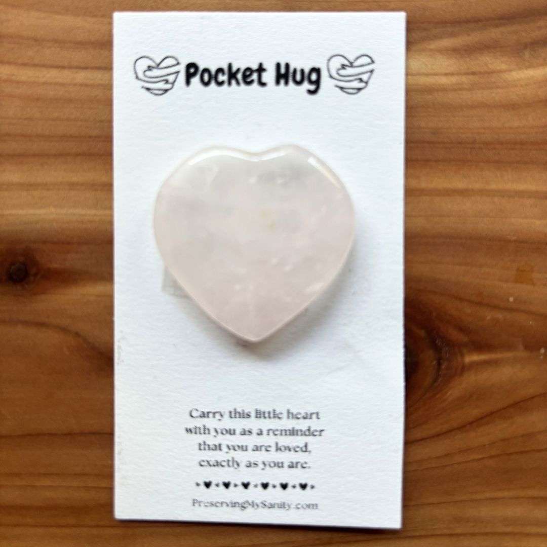 A polished heart-shaped rose quartz is mounted on a white "Pocket Hug" card and displayed on a warm cedar wood background. The card features the words "Pocket Hug" at the top with heart illustrations and a heartfelt message below: "Carry this little heart with you as a reminder that you are loved, exactly as you are." Small black hearts decorate the bottom above the Preserving My Sanity website, making this a thoughtful keepsake for encouragement, comfort, or gifting.