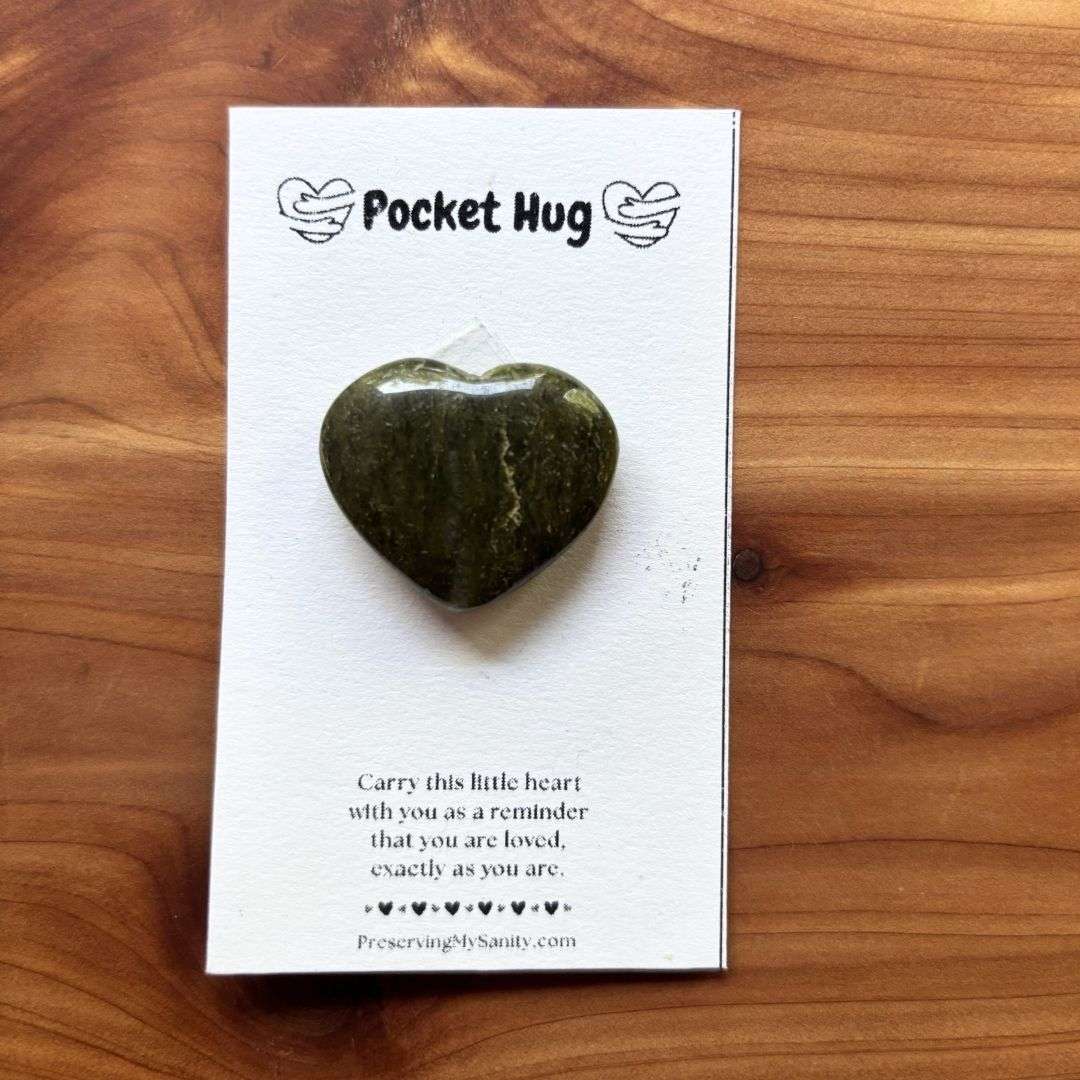 A polished heart-shaped vesuvianite is mounted on a white "Pocket Hug" card and displayed on a warm cedar wood background. The card features the words "Pocket Hug" at the top with heart illustrations and a heartfelt message below: "Carry this little heart with you as a reminder that you are loved, exactly as you are." Small black hearts decorate the bottom above the Preserving My Sanity website, making this a thoughtful keepsake for encouragement, comfort, or gifting.