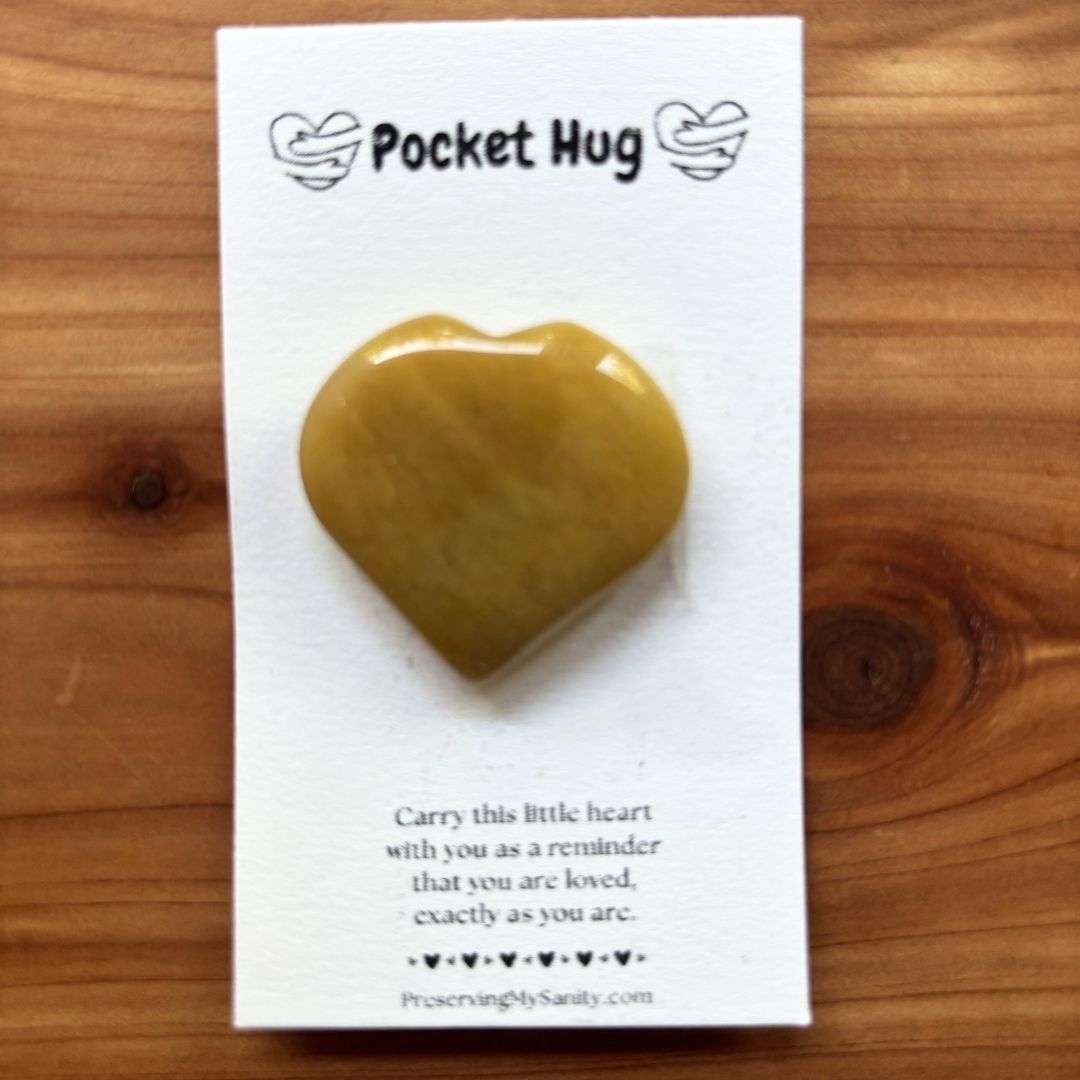 A polished heart-shaped yellow aventurine is mounted on a white "Pocket Hug" card and displayed on a warm cedar wood background. The card features the words "Pocket Hug" at the top with heart illustrations and a heartfelt message below: "Carry this little heart with you as a reminder that you are loved, exactly as you are." Small black hearts decorate the bottom above the Preserving My Sanity website, making this a thoughtful keepsake for encouragement, comfort, or gifting.