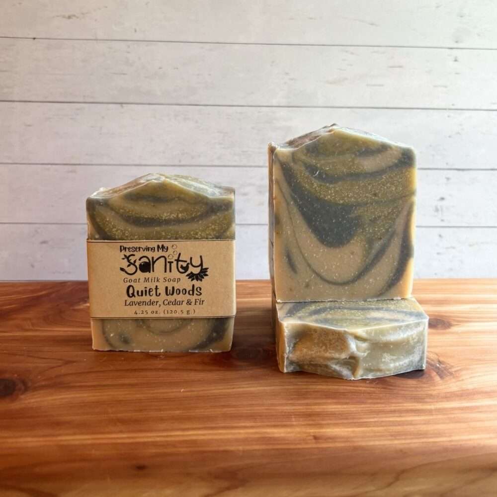 Three bars of Quiet Woods goat milk soap are displayed on a cedar bath board against a white shiplap background. One bar stands upright with the signature kraft paper label, one stands upright without a label to showcase its elegant marbled swirls of soft sage green, olive, and cream, and a third bar lies flat in front, revealing the textured top. Scented with lavender, cedar, and fir, this handcrafted soap features a calming woodland-inspired design that reflects its fresh, forest-like fragrance.