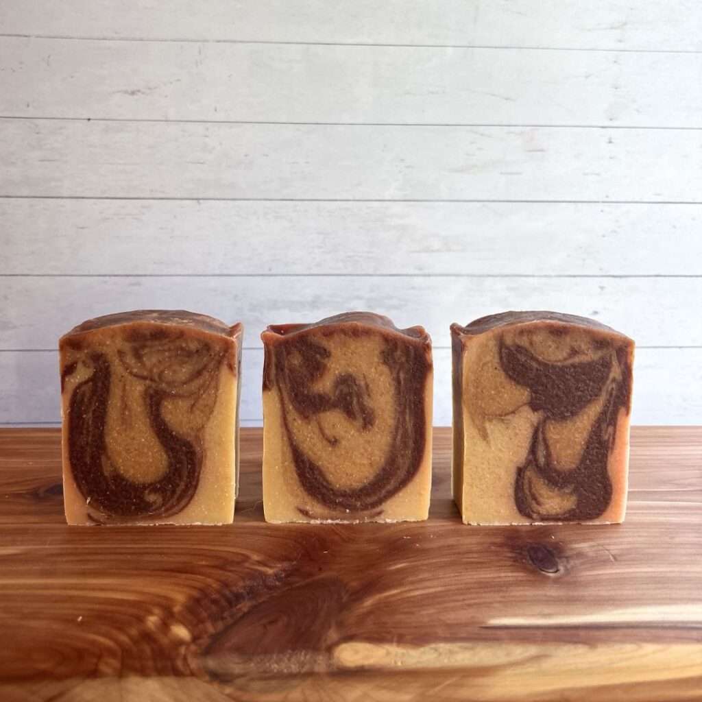 Three unwrapped bars of Rustic Radiance Goat Milk Soap are arranged in a neat row on a warm cedar wood bath board against a white shiplap background. Each handcrafted bar features a warm golden beige base with bold cocoa brown swirls that flow across the face of the soap, creating a unique marbled pattern on every bar.