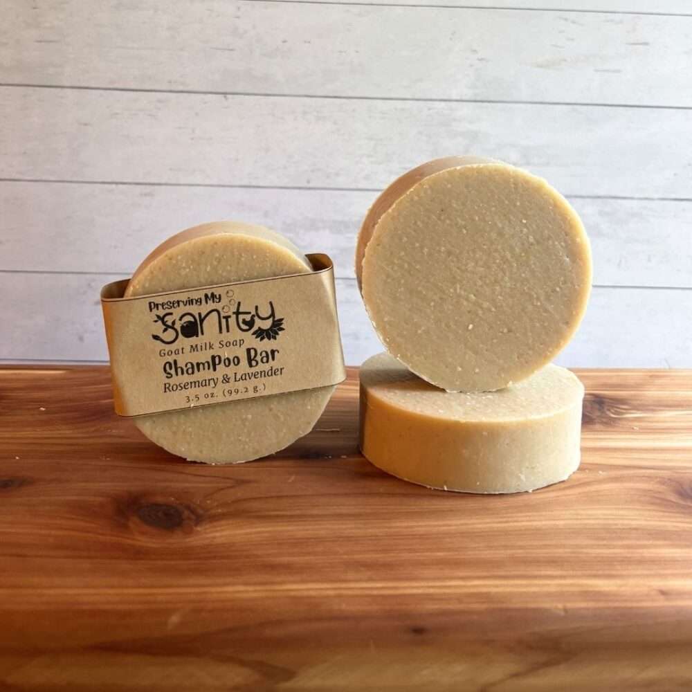 Three round Rosemary & Lavender shampoo bars are displayed on a warm cedar wood bath board against a white shiplap background. One bar is wrapped with the signature kraft paper label reading "Shampoo Bar," while a second bar stands upright to showcase its smooth, creamy surface. A third bar lies flat beneath it, highlighting the thick, solid design. The simple, natural appearance reflects this handcrafted shampoo bar scented with rosemary and lavender.