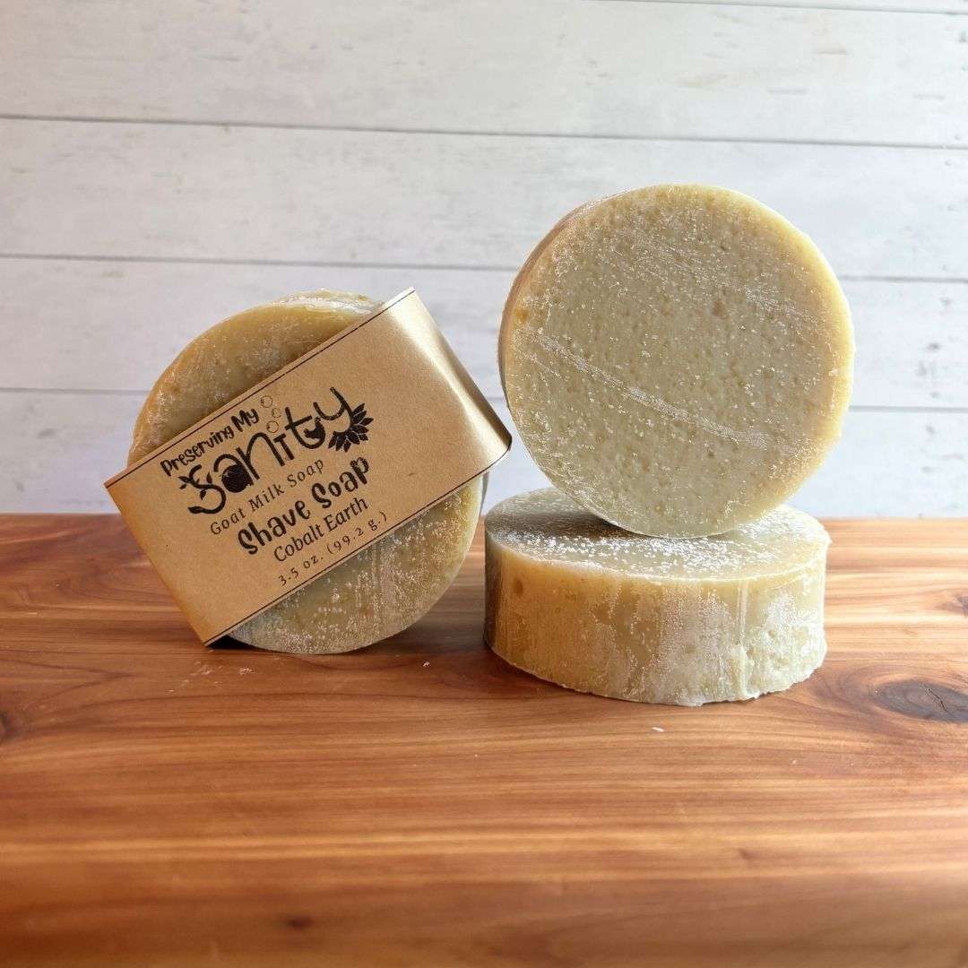 Three round Cobalt Earth shave soap bars are displayed on a warm cedar wood bath board against a white shiplap background. One bar stands upright with the signature kraft paper label reading "Shave Soap - Cobalt Earth," a second unwrapped bar stands upright to showcase its smooth, creamy ivory surface, and a third bar lies flat in front, revealing the thick, solid design. Scented with cedarwood, bergamot, and patchouli essential oils, this handcrafted shave soap has a clean, natural appearance.