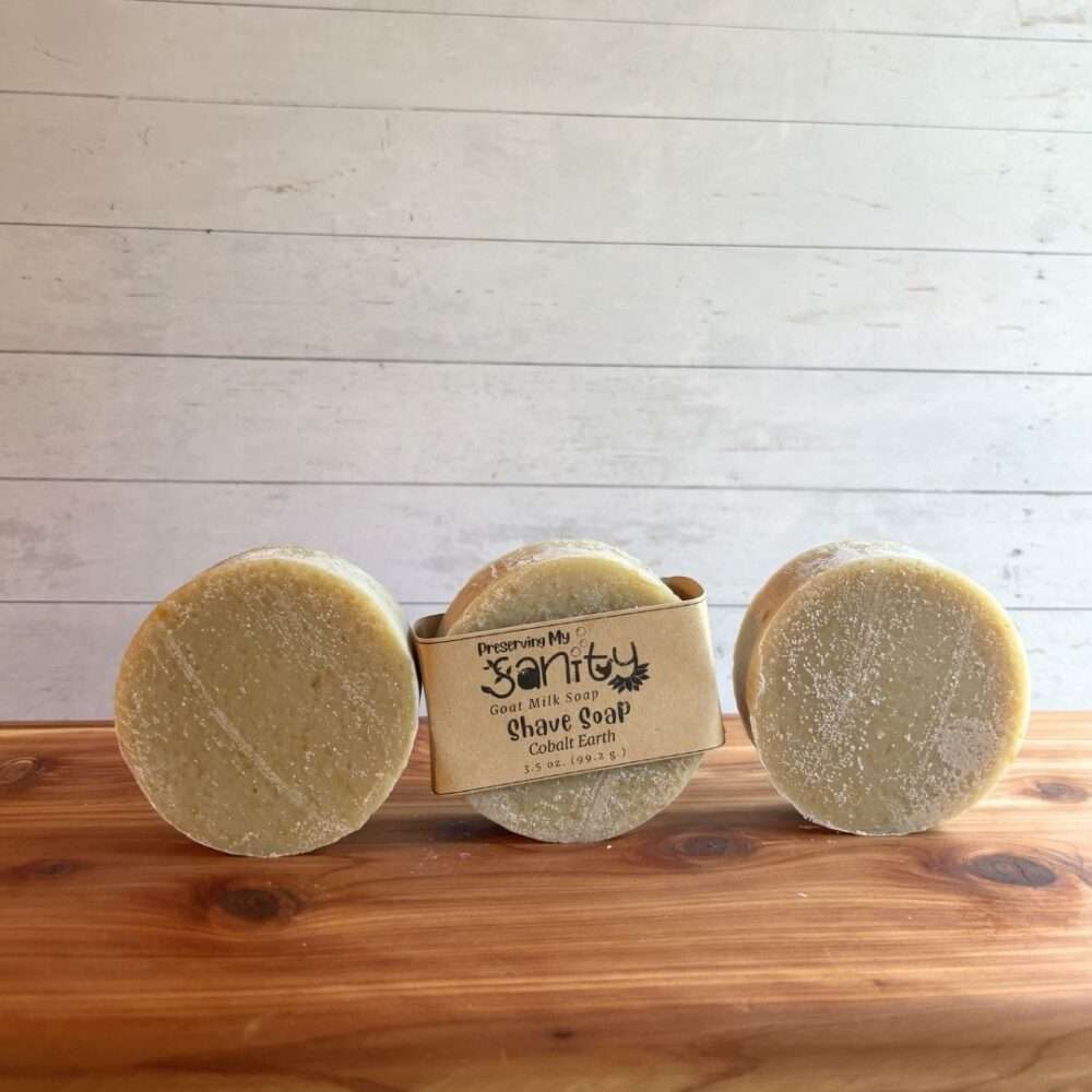 Three round Cobalt Earth shave soap bars are arranged in a row on a warm cedar wood bath board against a white shiplap background. The center bar is wrapped with the signature kraft paper label reading "Shave Soap - Cobalt Earth," while the bars on the left and right are unwrapped, showcasing their smooth, creamy ivory surface and round shape.