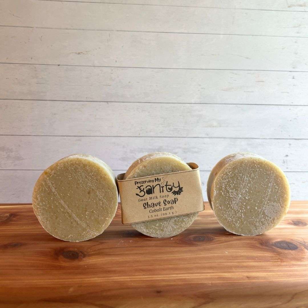 Three round Cobalt Earth shave soap bars are arranged in a row on a warm cedar wood bath board against a white shiplap background. The center bar is wrapped with the signature kraft paper label reading "Shave Soap - Cobalt Earth," while the bars on the left and right are unwrapped, showcasing their smooth, creamy ivory surface and round shape.