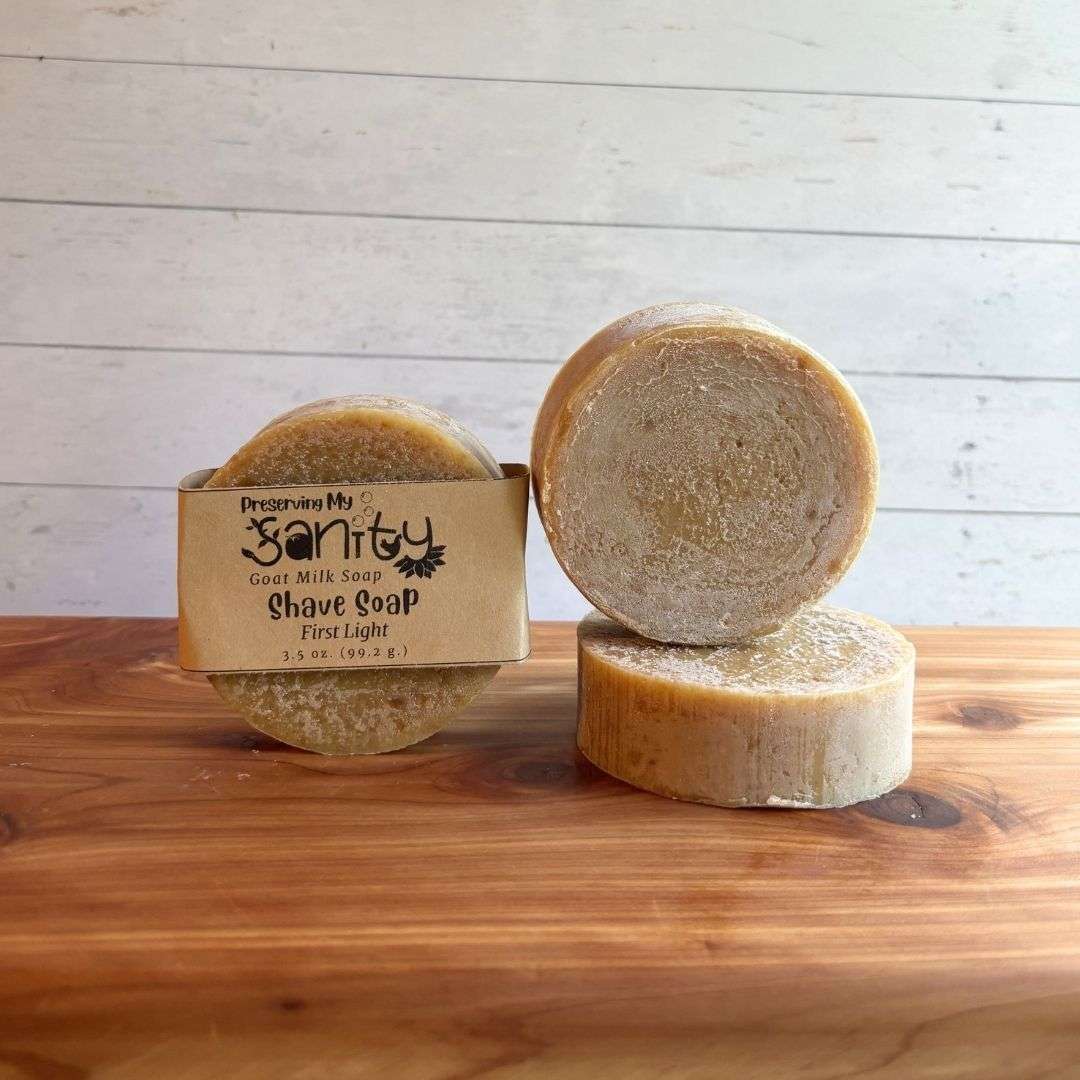 Three round First Light shave soap bars are displayed on a warm cedar wood bath board against a white shiplap background. One bar stands upright with the signature kraft paper label reading "Shave Soap - First Light." A second unwrapped bar stands upright beside it, showcasing its smooth, naturally tan surface, while a third bar lies flat in front to display the top of the soap. Scented with litsea cubeba and orange essential oils and naturally colored with spinach powder, this handcrafted shave soap has a clean, simple appearance.