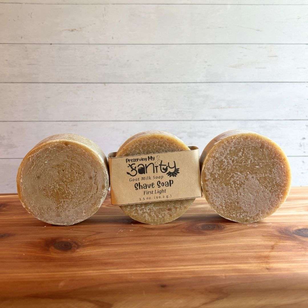 Three round First Light shave soap bars are arranged in a neat row on a warm cedar wood bath board against a white shiplap background. The center bar is wrapped with the signature kraft paper label reading "Shave Soap - First Light," while the matching bars on either side are unwrapped, showcasing their smooth, naturally tan color and round shape. Made with litsea cubeba and orange essential oils and naturally colored with spinach powder, the handcrafted bars have a clean, minimalist presentation.