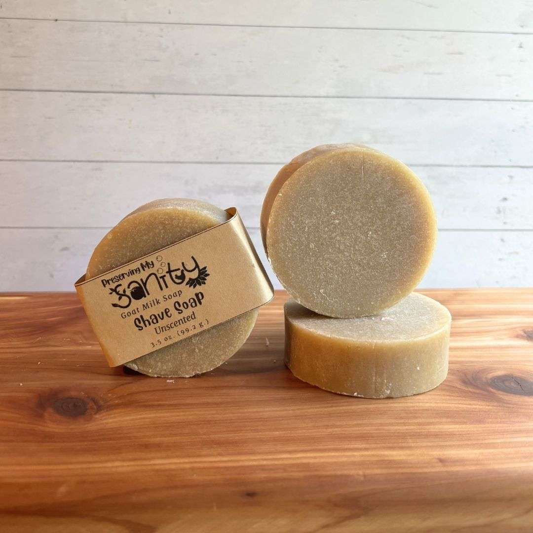 Three round unscented shave soap bars are displayed on a warm cedar wood bath board against a white shiplap background. One bar stands upright with the signature kraft paper label reading "Shave Soap - Unscented," a second unwrapped bar stands upright to showcase its smooth, creamy ivory surface, and a third bar lies flat in front, revealing the top of the soap. Free of essential oils and colorants, this handcrafted shave soap has a clean, simple appearance that highlights its natural, fragrance-free formula.