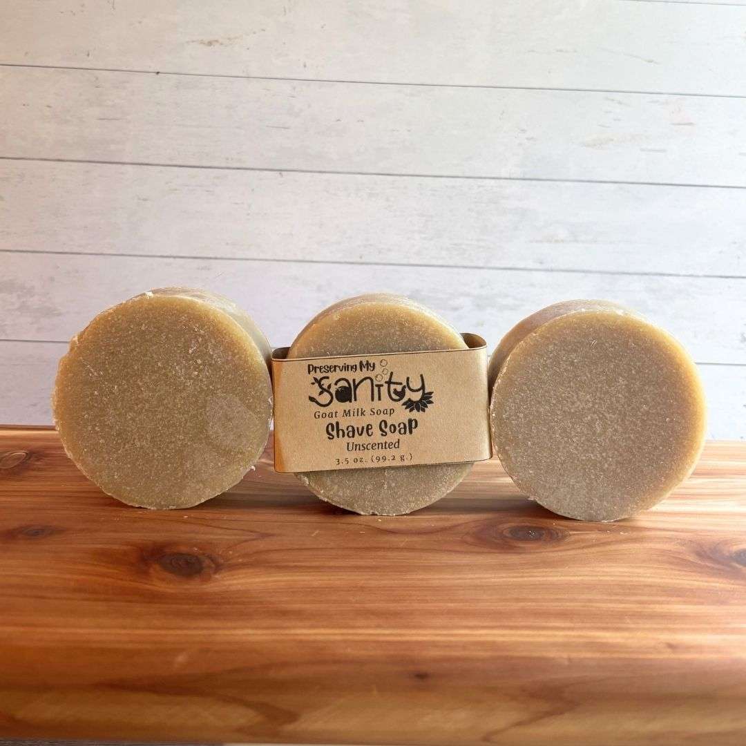 Three round unscented shave soap bars are arranged in a straight row on a warm cedar wood bath board against a white shiplap background. The center bar is wrapped in the signature kraft paper label reading "Shave Soap - Unscented," while the bars on either side are unwrapped, showcasing their smooth, creamy ivory color and simple, natural appearance. Made without essential oils or colorants, this fragrance-free shave soap highlights its clean, minimalist formulation.