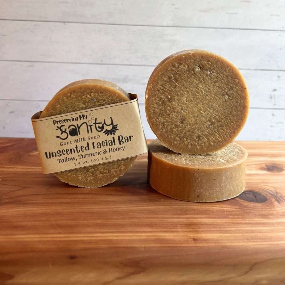 One round bar of Unscented Turmeric & Honey Facial Soap stands upright with the kraft paper label visible, beside a second unwrapped bar standing upright on top of a third bar laid flat to display its thickness. The soap has a naturally warm golden color from turmeric and a smooth, slightly textured surface. Displayed on a warm cedar wood bath board against a white shiplap background, the fragrance-free facial soap highlights its simple, handcrafted design made with goat milk, raw honey, and nourishing oils.