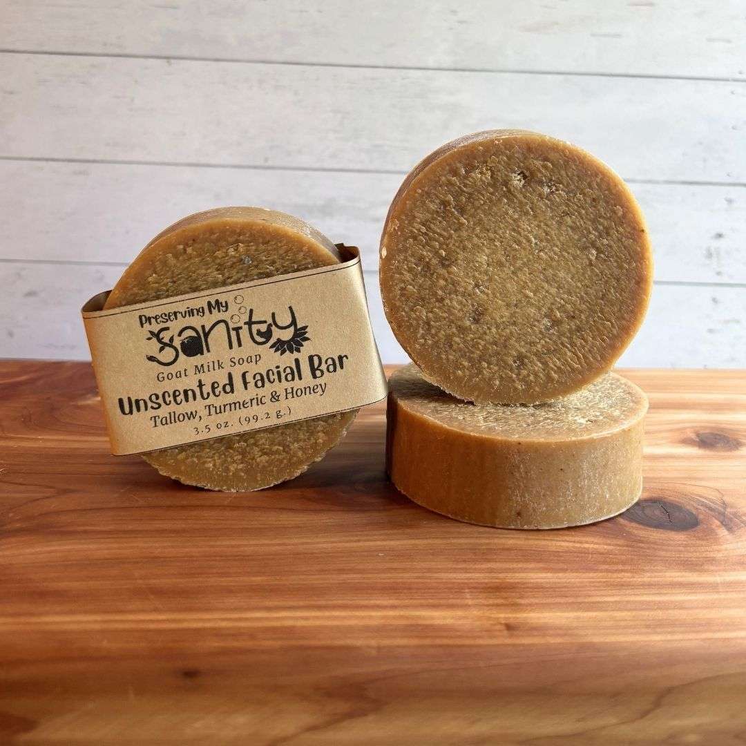 One round bar of Unscented Turmeric & Honey Facial Soap stands upright with the kraft paper label visible, beside a second unwrapped bar standing upright on top of a third bar laid flat to display its thickness. The soap has a naturally warm golden color from turmeric and a smooth, slightly textured surface. Displayed on a warm cedar wood bath board against a white shiplap background, the fragrance-free facial soap highlights its simple, handcrafted design made with goat milk, raw honey, and nourishing oils.