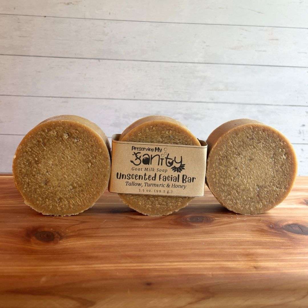Three round bars of Unscented Turmeric & Honey Facial Soap are arranged side by side in a row on a warm cedar wood bath board against a white shiplap background. The center bar is wrapped with the kraft paper label, while the bars on the left and right are unwrapped, showcasing the soap's naturally warm golden color, smooth surface, and handcrafted finish.