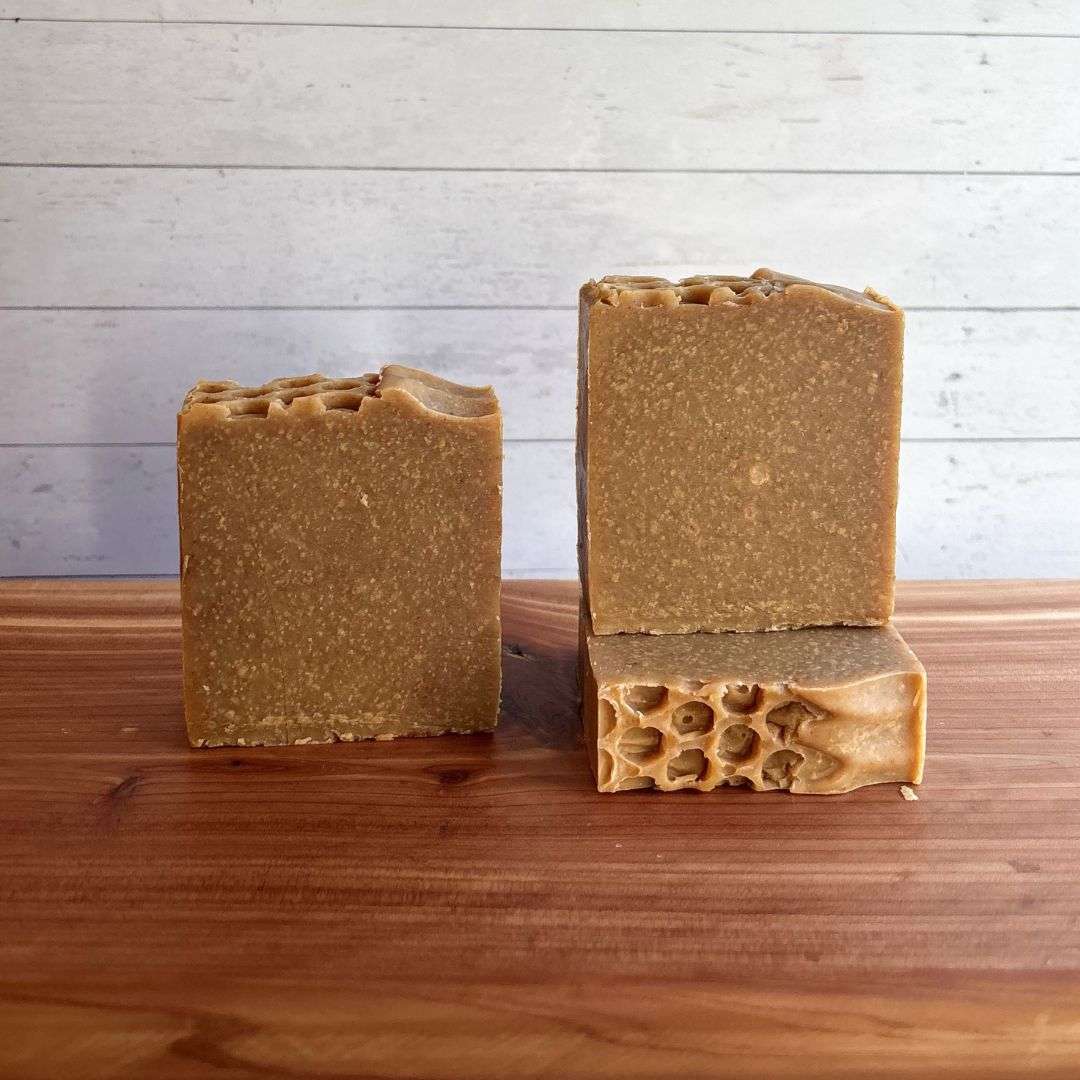 Three unlabelled bars of Turmeric, Oats & Honey Goat Milk Soap are displayed on a warm cedar wood bath board against a white shiplap background. Two bars stand upright side by side, showcasing their warm golden tan color with fine natural speckles throughout and rustic honeycomb-textured tops. A third bar lies flat beneath one of the upright bars, revealing the decorative honeycomb pattern across the top surface.