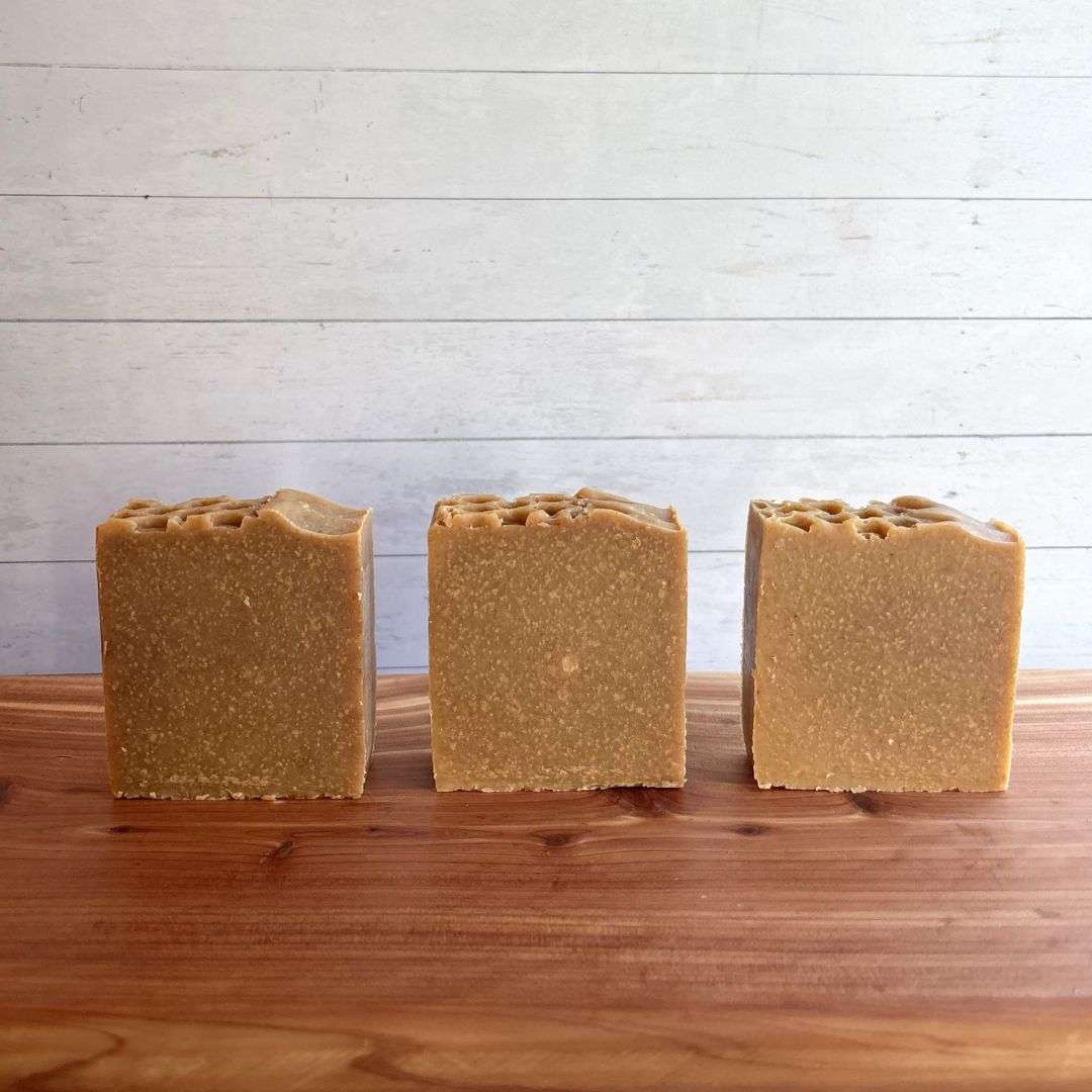 Three unlabelled bars of Turmeric, Oats & Honey Goat Milk Soap are arranged in a neat row on a warm cedar wood bath board against a white shiplap background. Each handcrafted bar has a warm golden tan color with fine natural speckles throughout, giving it a wholesome, rustic appearance. The tops feature a distinctive honeycomb-inspired texture that highlights the artisan craftsmanship.