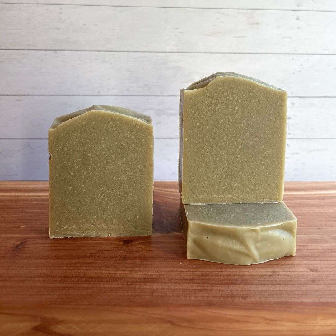 Three unlabelled bars of Unscented Avocado Goat Milk Soap are displayed on a warm cedar wood bath board against a white shiplap background. Two bars stand upright side by side, showcasing their soft natural green color, smooth sides, and gently sculpted artisan tops. A third bar lies flat beneath one of the upright bars, revealing the smooth top surface and creamy texture.