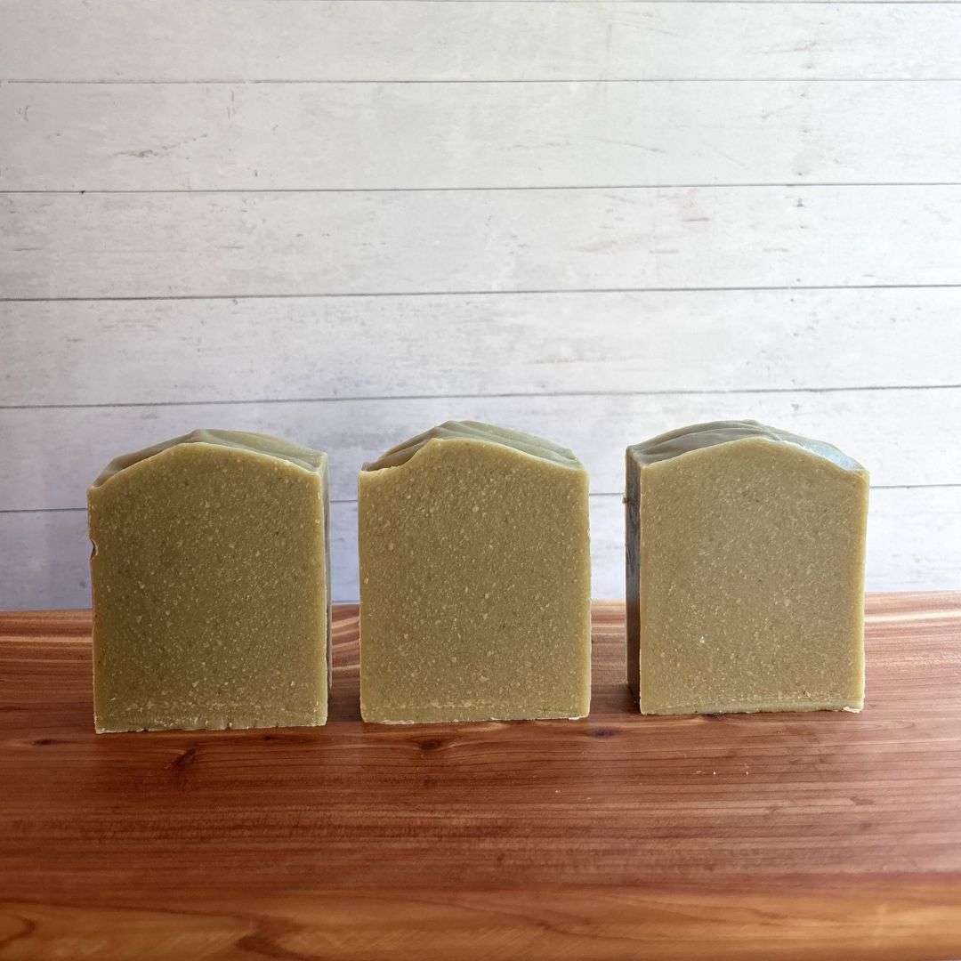 Three unlabelled bars of Unscented Avocado Goat Milk Soap are arranged in a neat row on a warm cedar wood bath board against a white shiplap background. Each handcrafted bar has a soft natural green color with a smooth, minimalist design and gently sculpted tops.