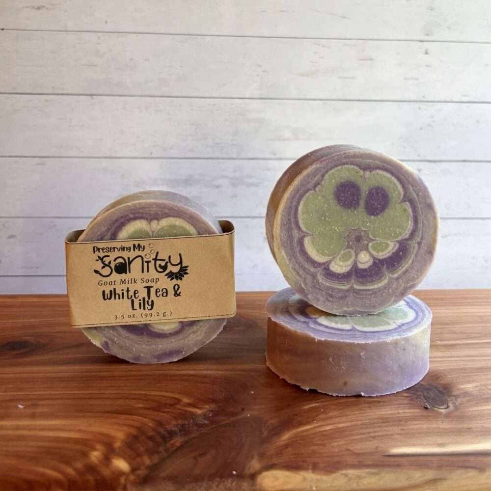 Three round bars of White Tea & Lily Goat Milk Soap are displayed on a warm cedar wood bath board against a white shiplap background. One bar stands upright with the signature kraft paper label reading "White Tea & Lily." A second unwrapped bar stands upright beside it, showcasing the intricate floral-inspired design in soft lavender, sage green, and cream. A third unwrapped bar lies flat in front, displaying the top ad the soap’s thickness.