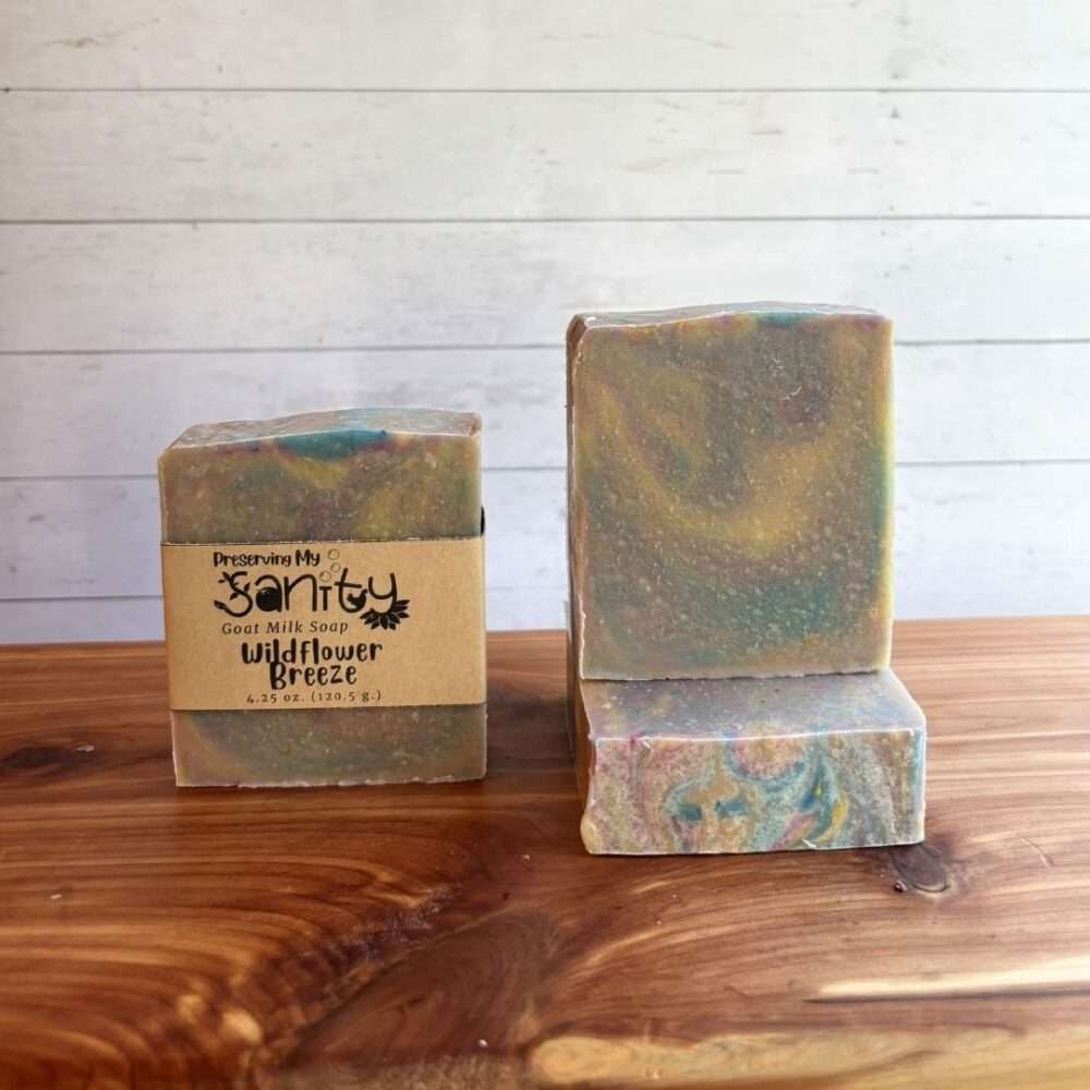Three bars of Wildflower Breeze Goat Milk Soap are displayed on a warm cedar wood bath board against a white shiplap background. One bar stands upright with the signature kraft paper label reading "Wildflower Breeze." A second unwrapped bar stands upright beside it, showcasing soft swirls of muted sage green, dusty blue, blush, and warm beige. A third unwrapped bar lies flat in front, displaying the colorful swirl pattern across the top.
