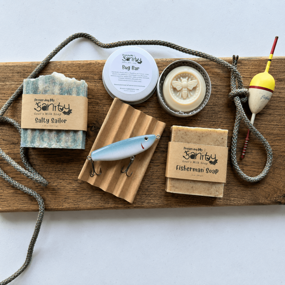 Fisherman's Bundle arranged on a rustic wooden board with a white background. The gift set includes a bar of Salty Sailor goat milk soap, a 3-ounce bar of Fisherman's Soap wrapped in a kraft label, a tin of Bug Lotion Bar with the lid removed to reveal the bee design, a natural wood soap dish topped with a handmade blue-and-white fishing lure, and a vintage fishing bobber. A length of rope is draped around the display, highlighting the fishing theme of this handcrafted Minnesota gift set.